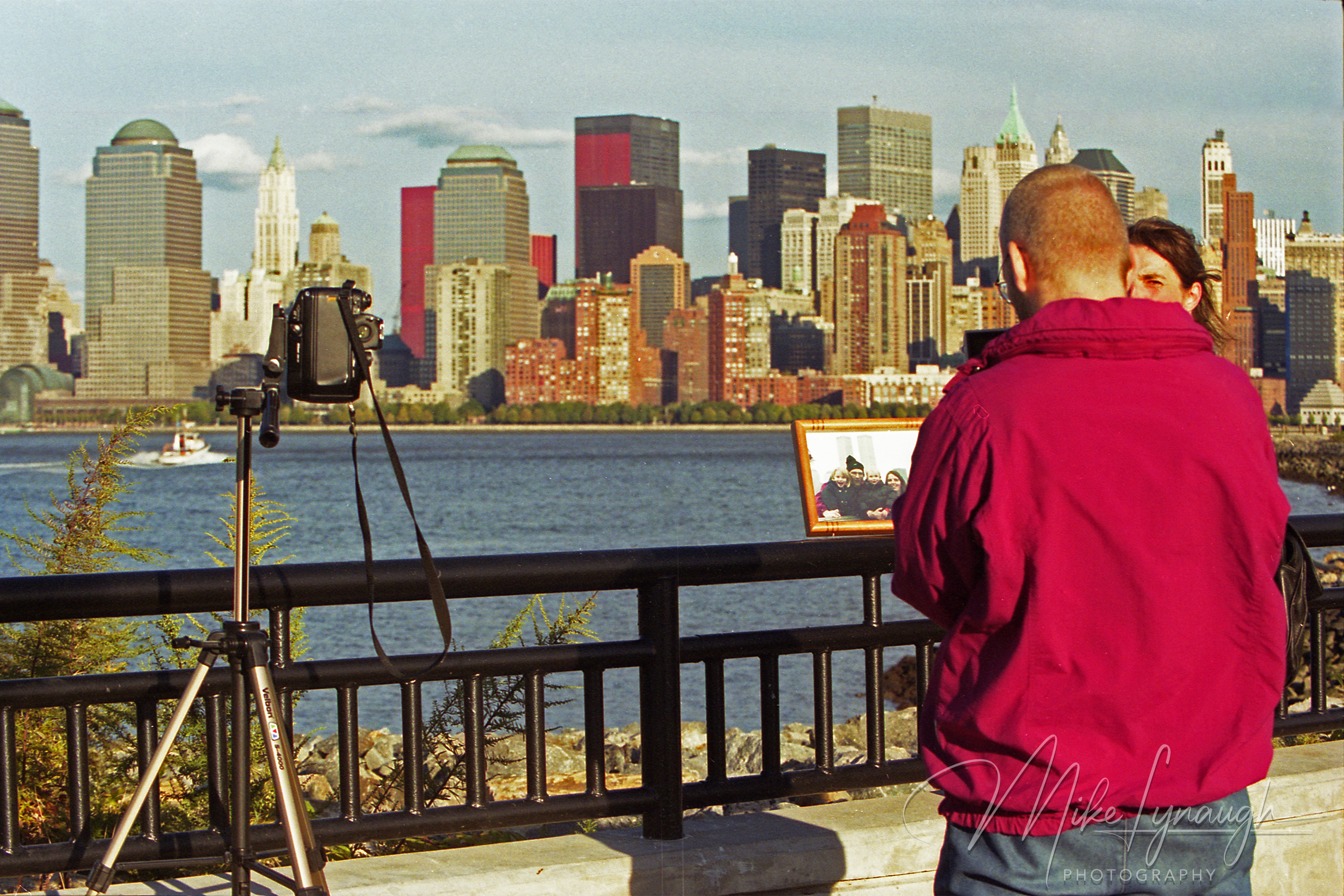 A man in a magenta jacket holding a framed photo of loved ones against the Lower Manhattan skyline from a Jersey City waterfront promenade in October 2001.