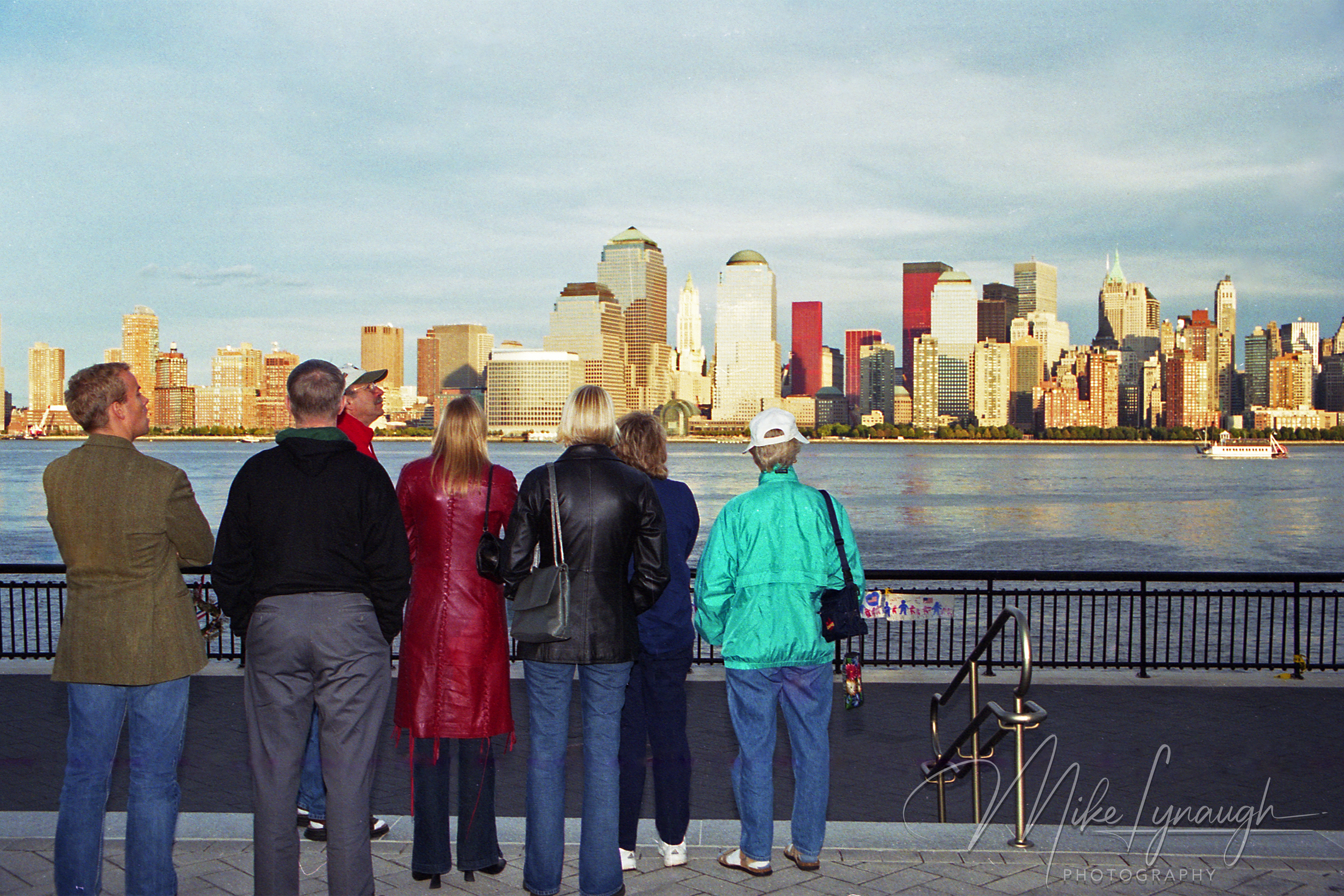 A group of people viewed from behind standing along a waterfront promenade railing, gazing across the water at the Lower Manhattan skyline in October 2001."