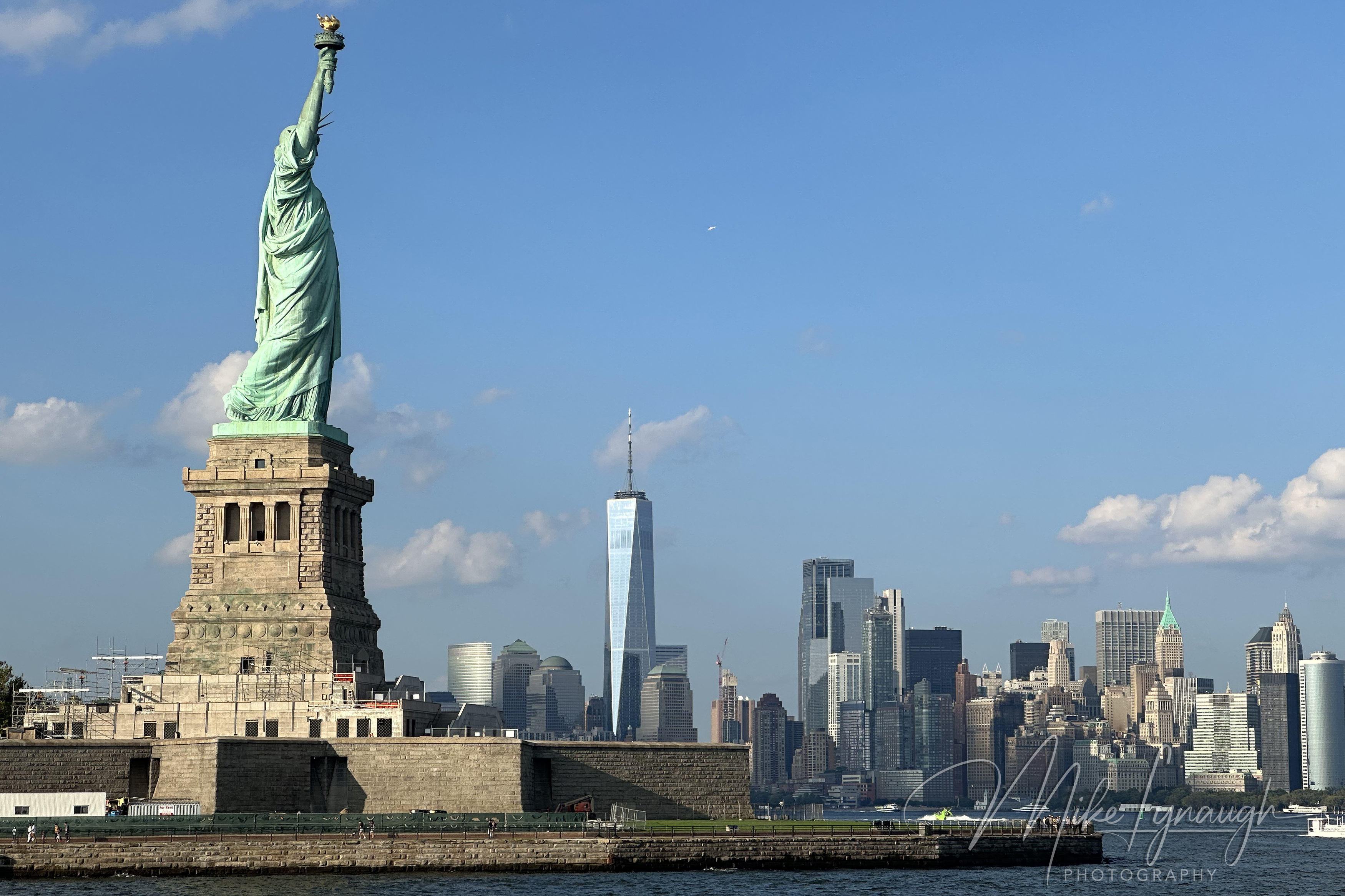 A wide harbor view showing the Statue of Liberty on the left with the completed One World Trade Center and the Lower Manhattan skyline in the background.