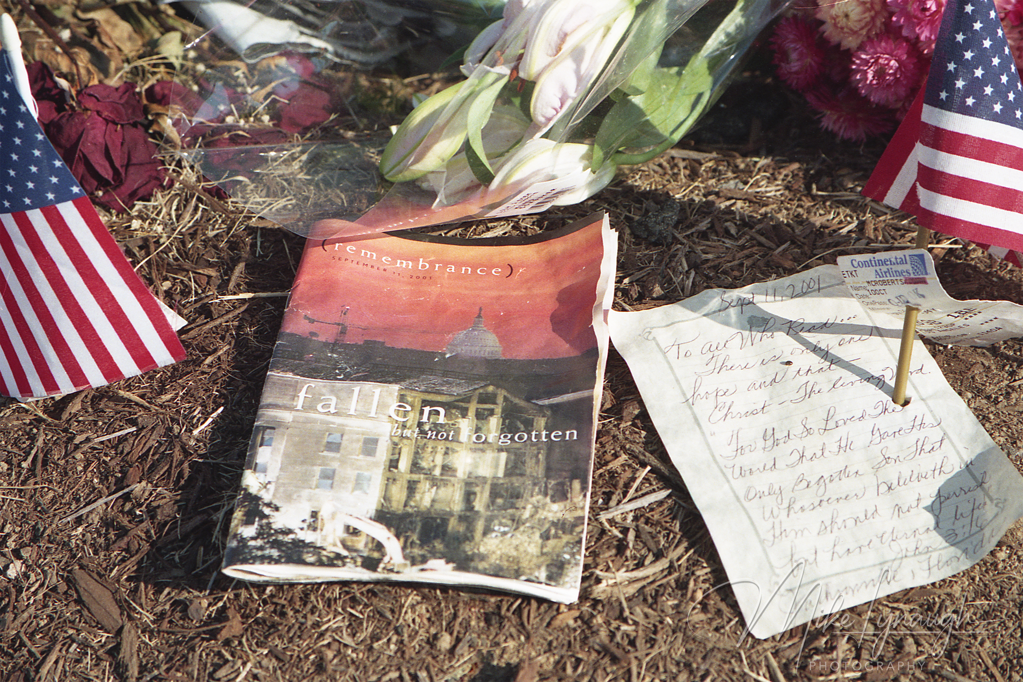 A close-up of a remembrance booklet reading fallen but not forgotten, small American flags, a handwritten Bible verse, and an airline ticket stub on the ground