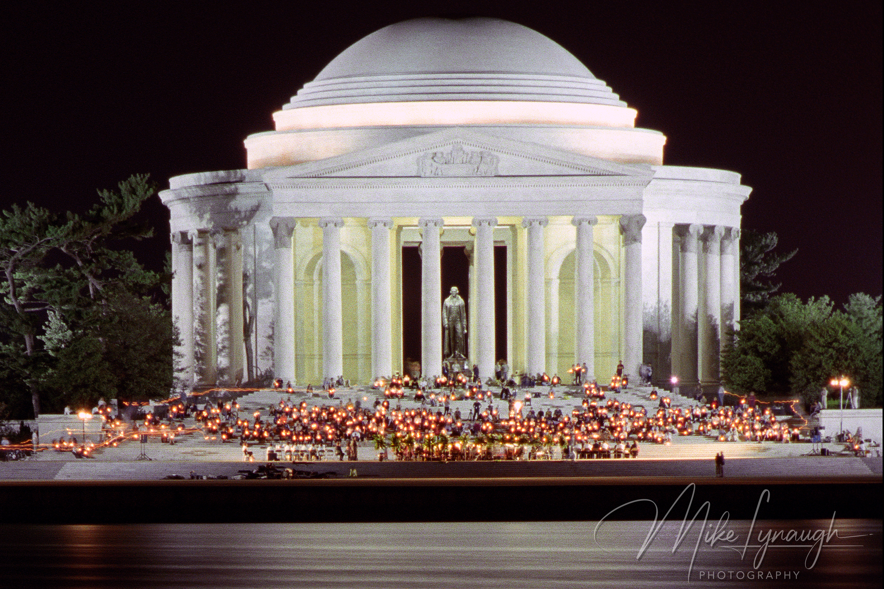 A night long-exposure photograph of the illuminated Jefferson Memorial across the Tidal Basin with crowds holding candles on the marble steps