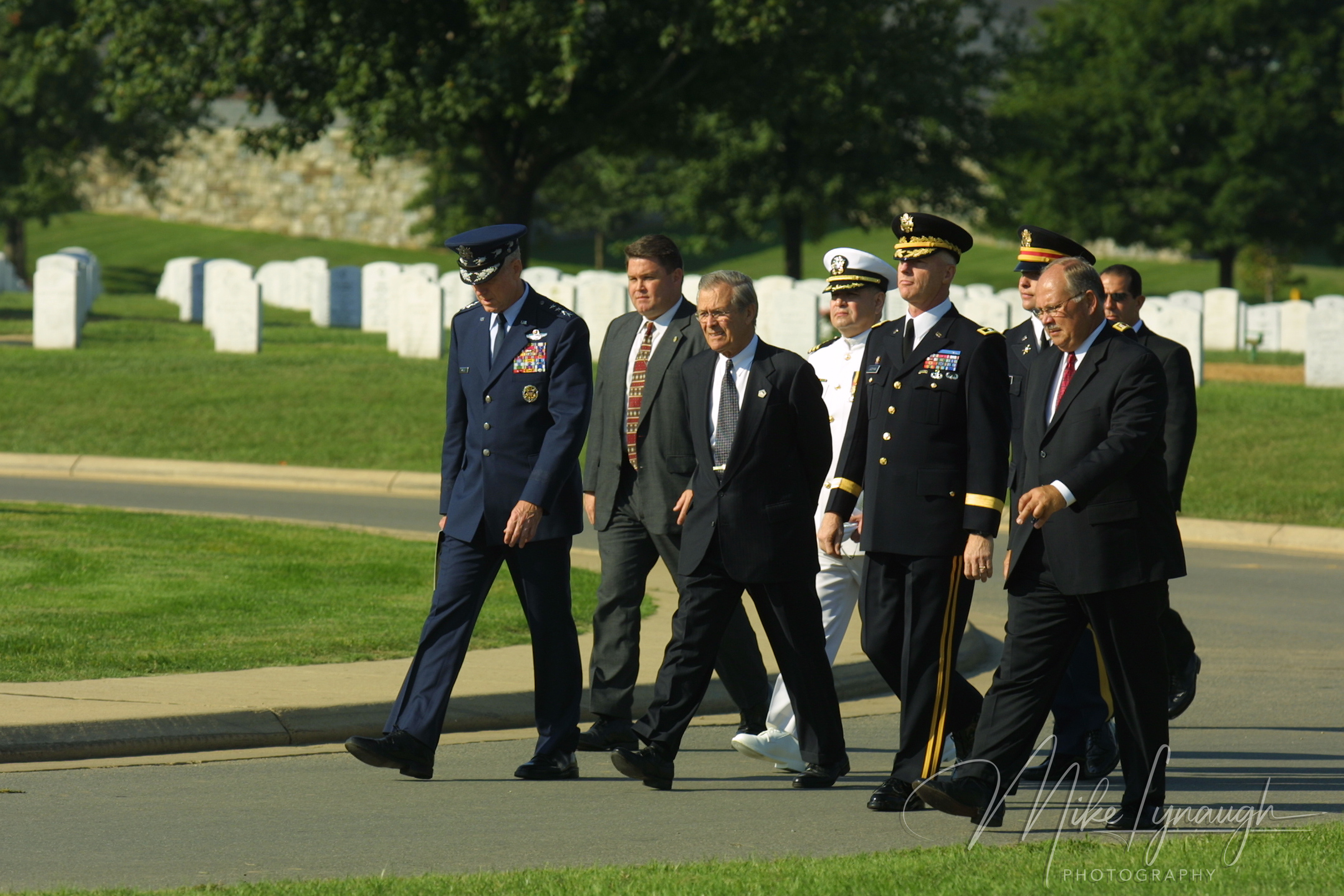 Secretary of Defense Donald Rumsfeld and military officers in uniform walking along a paved road in Arlington National Cemetery past rows of white headstones