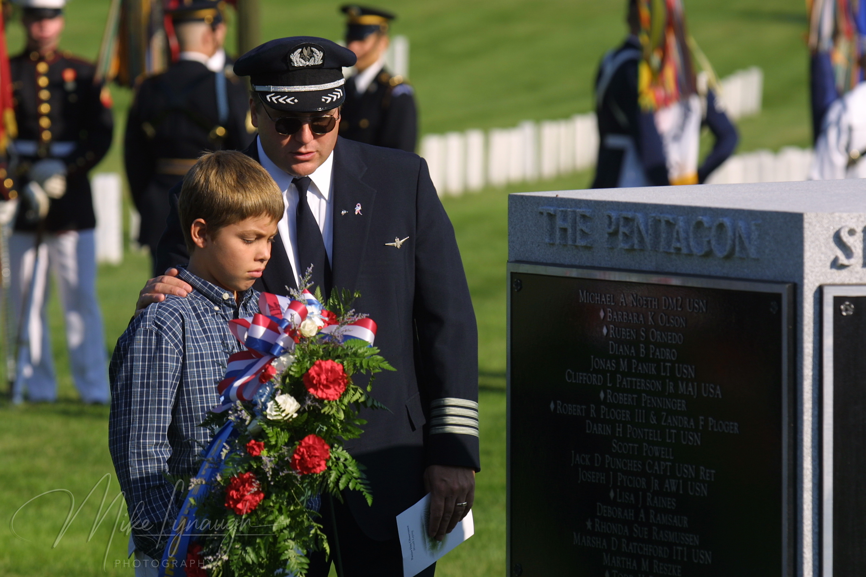 American Airlines Captain James C. Condes in uniform with sunglasses resting his hand on his son Christopher's shoulder in front of a floral wreath and the granite Pentagon 9/11 memorial marker at Arlington National Cemetery