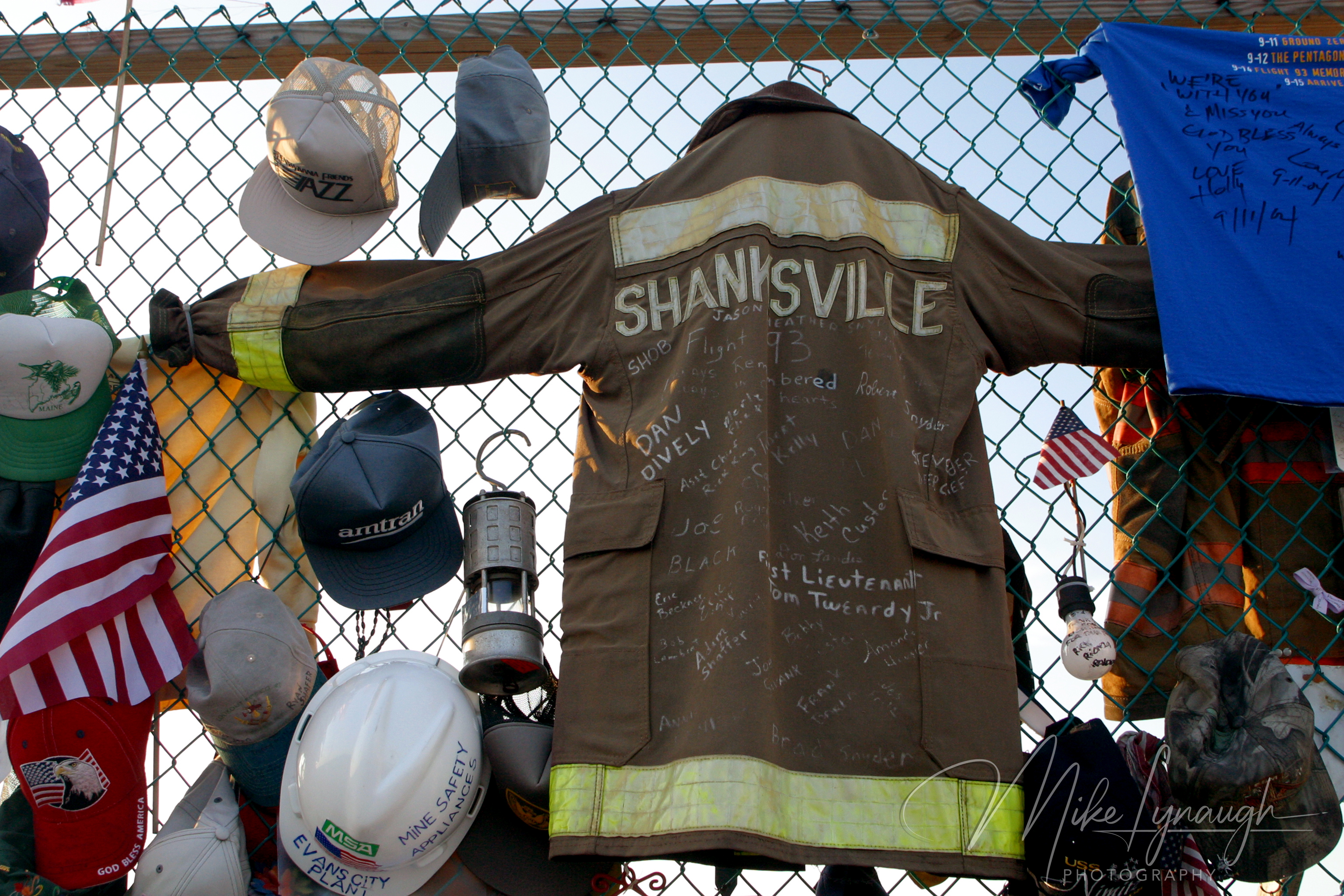 A close-up of a brown Shanksville firefighter jacket covered in handwritten signatures hanging on a wire fence surrounded by baseball caps, a white hard hat, a lantern, and an American flag.