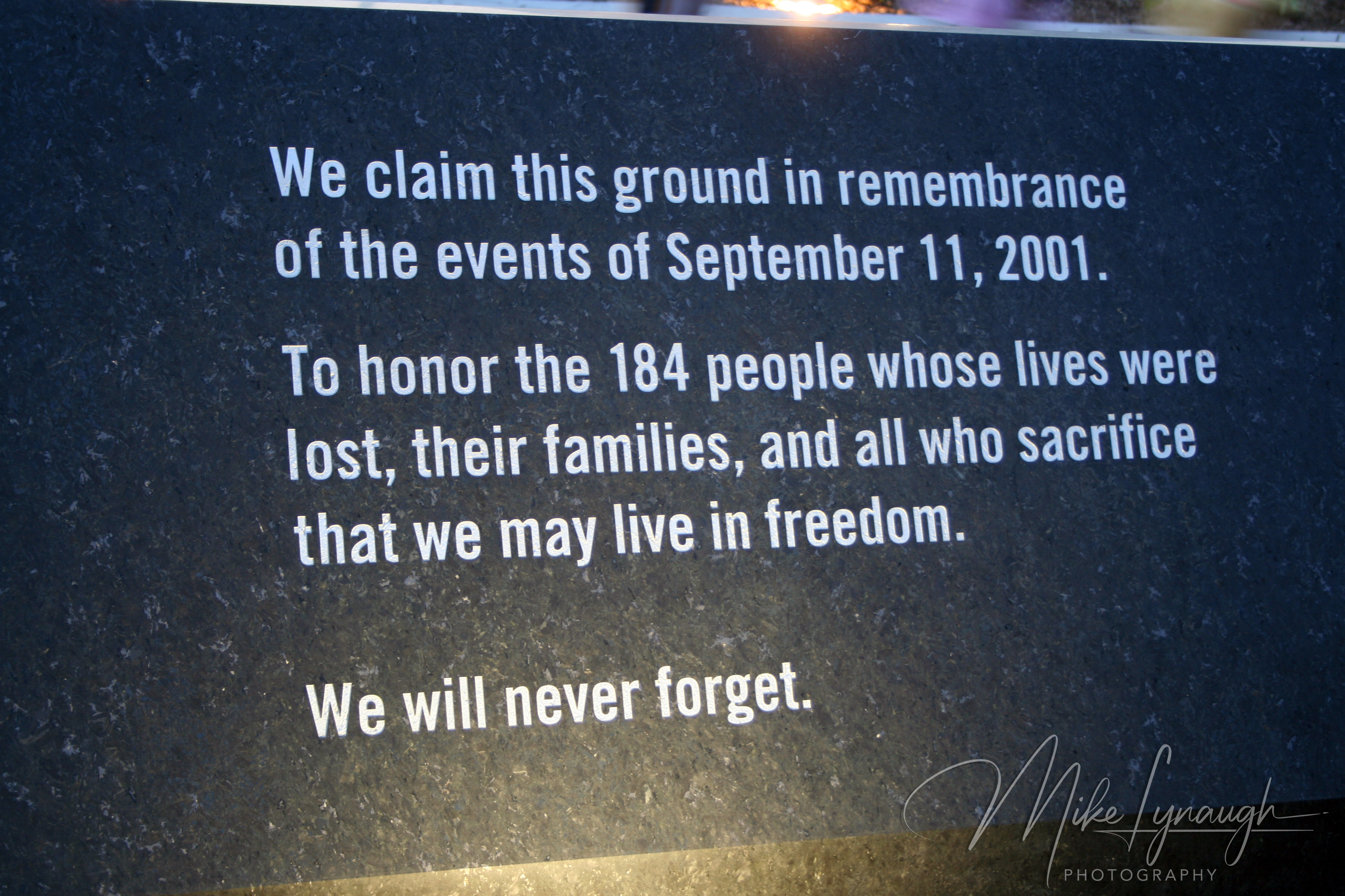 A close-up of an engraved black granite stone plaque reading 'We claim this ground in remembrance of the events of September 11, 2001. To honor the 184 people whose lives were lost, their families, and all who sacrifice that we may live in freedom. We will never forget.'
