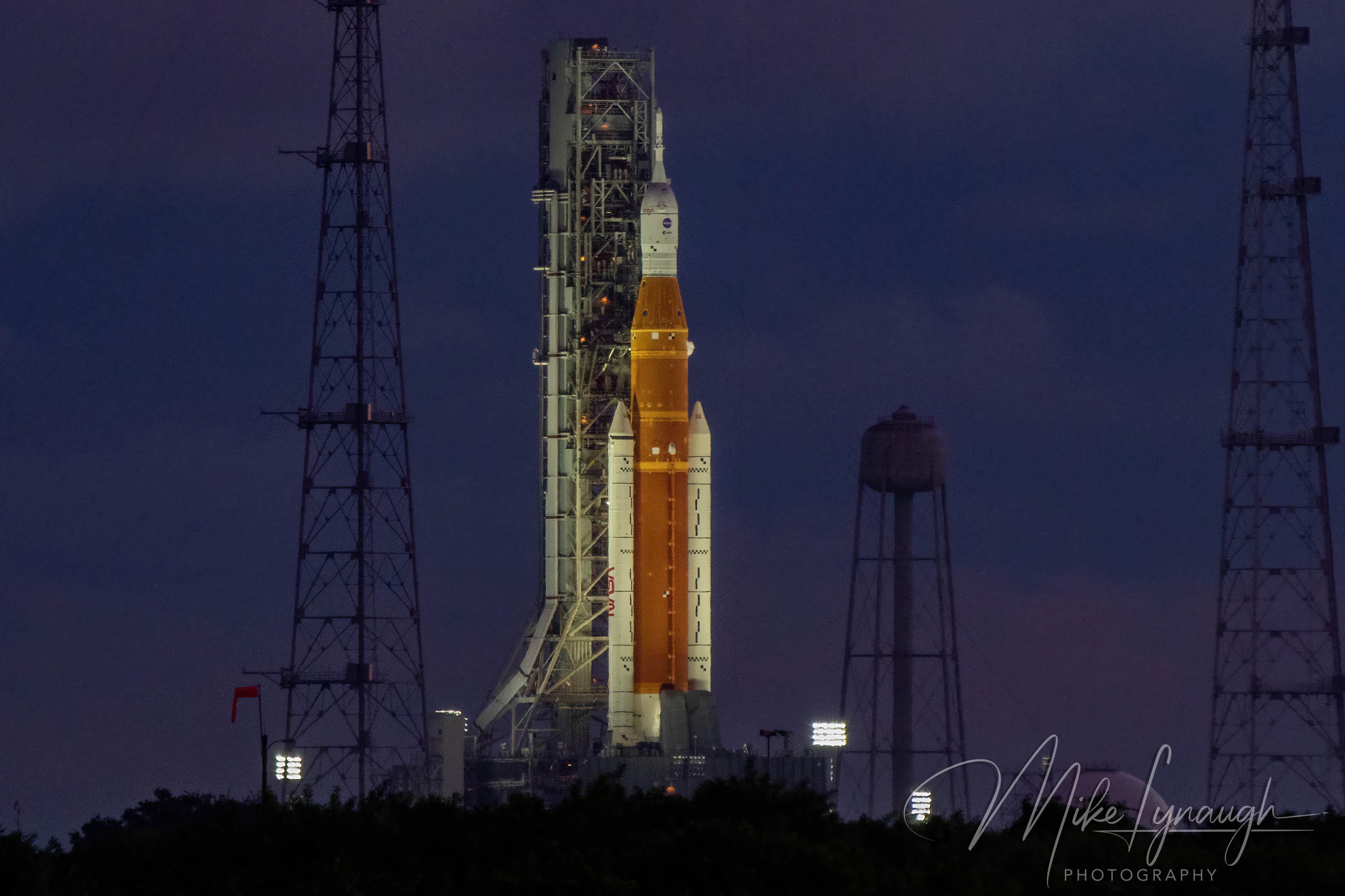 A horizontal telephoto photograph capturing NASA’s Space Launch System (SLS) rocket on Launch Pad 39B against a deep indigo dawn sky.