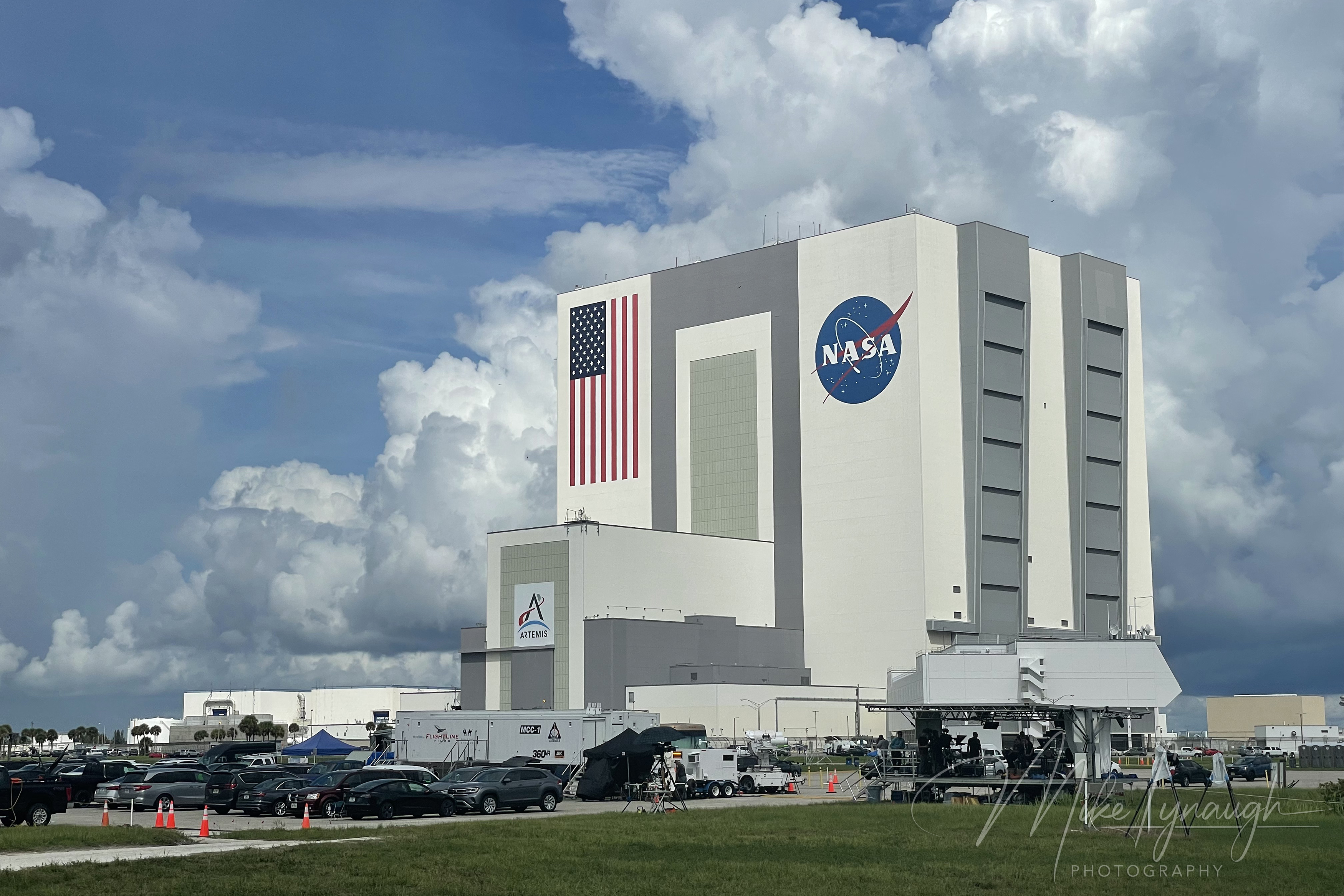 A wide three-quarter photograph of the 525-foot Vehicle Assembly Building (VAB) towering over the NASA Press Site lawn.
