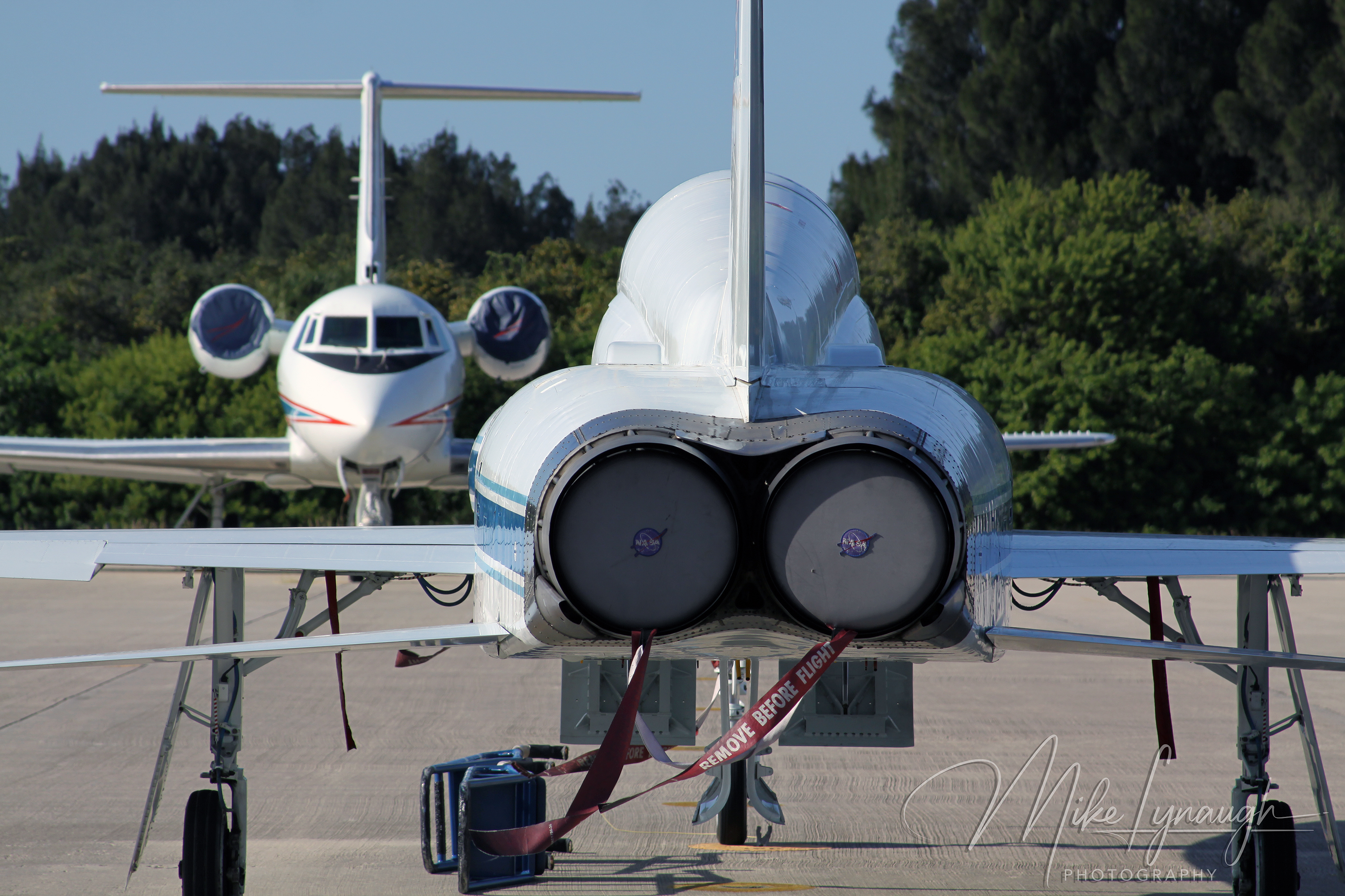 A horizontal outdoor telephoto photograph capturing the rear perspective of a NASA Northrop T-38N Talon jet lined up in front of a NASA Gulfstream II transport aircraft on the apron at the Shuttle Landing Facility (SLF).