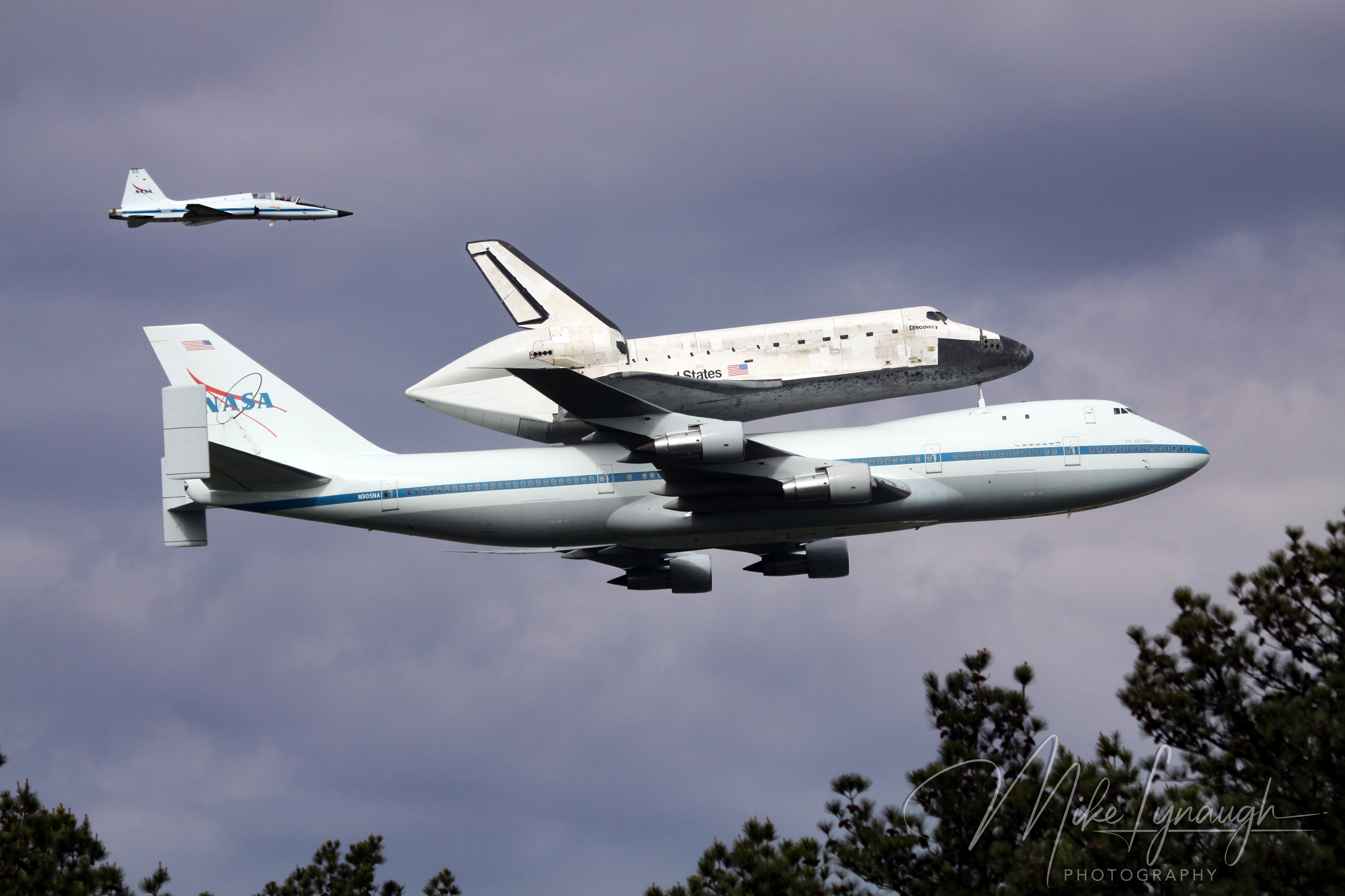 A wide-angle formation photograph capturing NASA’s Boeing 747 Shuttle Carrier Aircraft (SCA 905) ferrying Space Shuttle Discovery (OV-103), escorted by a NASA T-38 Talon chase plane.