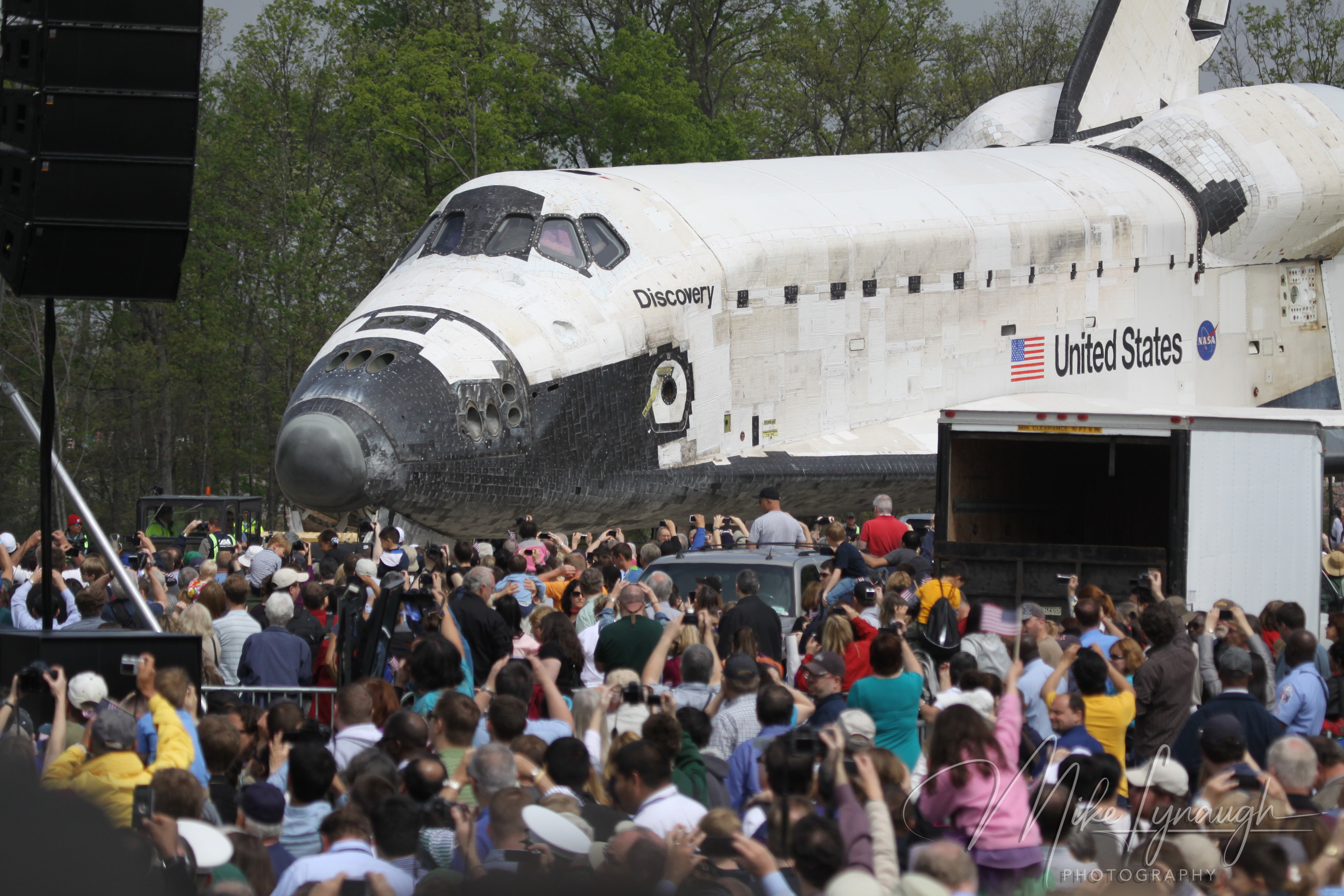 A telephoto profile photograph capturing Space Shuttle Discovery (OV-103) moving past a large crowd of spectators and media at the Steven F.