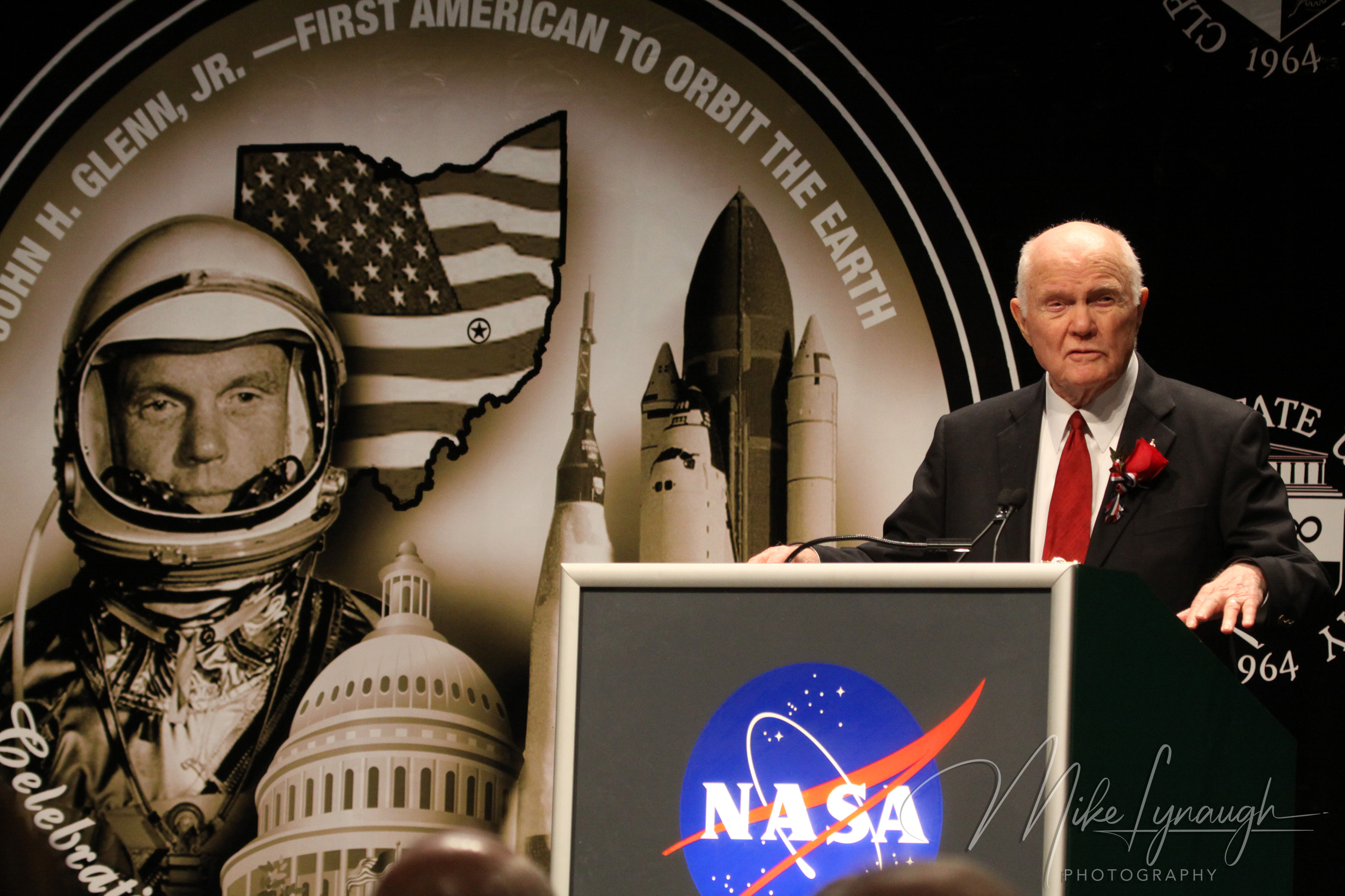 A medium landscape photograph capturing legendary astronaut and U.S. Senator John Glenn standing at a NASA-branded podium as he delivers his speech.