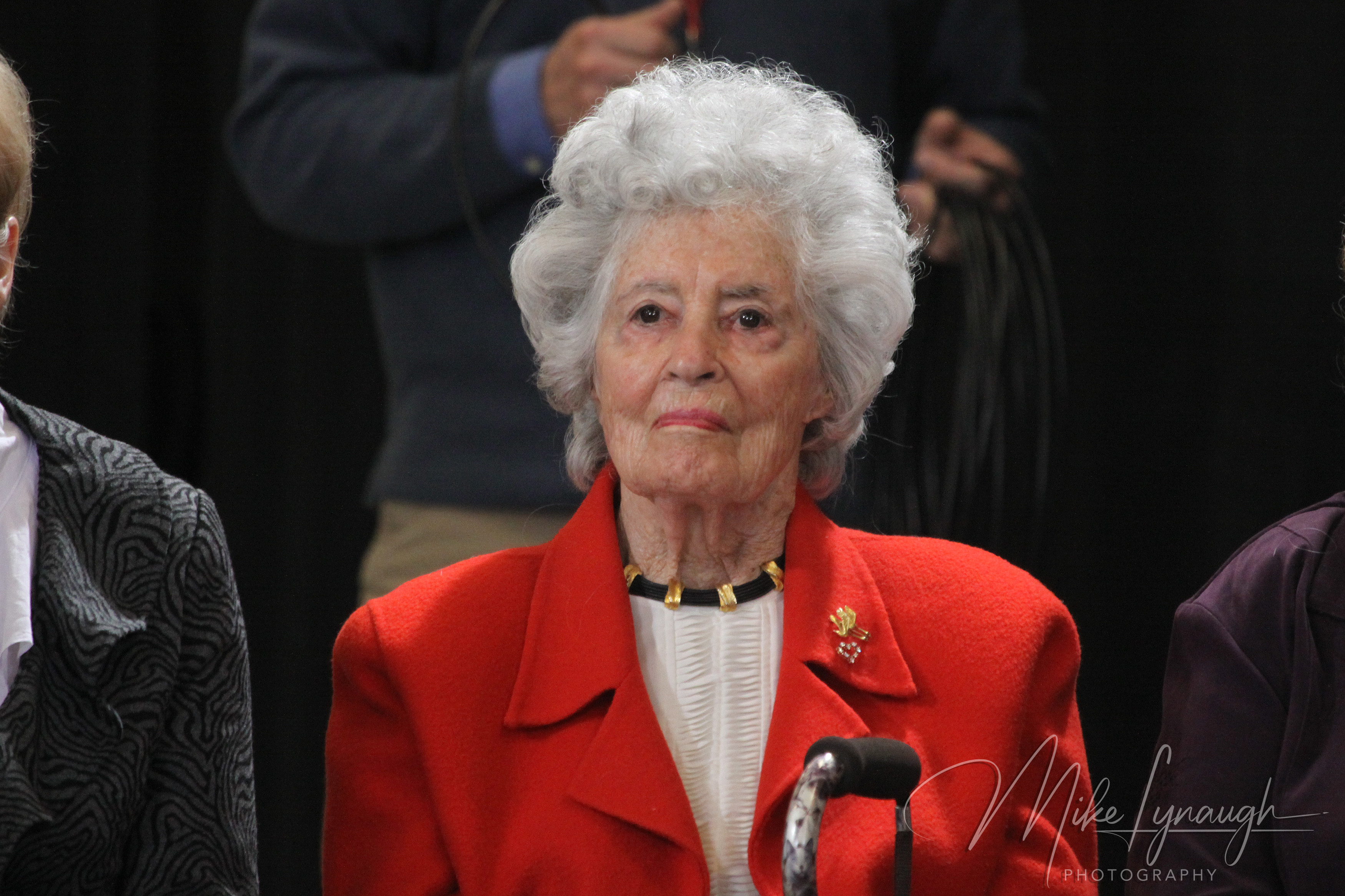 A centered medium-close portrait photograph capturing Annie Glenn, advocate and wife of astronaut Senator John Glenn, wearing a vibrant red jacket and gold necklace while seated in the audience.