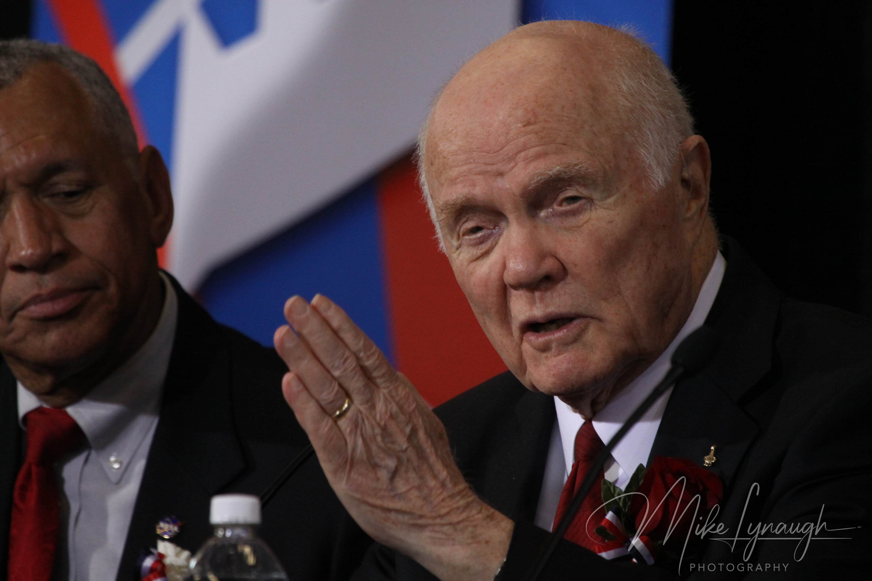 A close-up photograph capturing legendary astronaut and former U.S. Senator John Glenn gesturing with his hand near the microphone as he speaks to attendees.