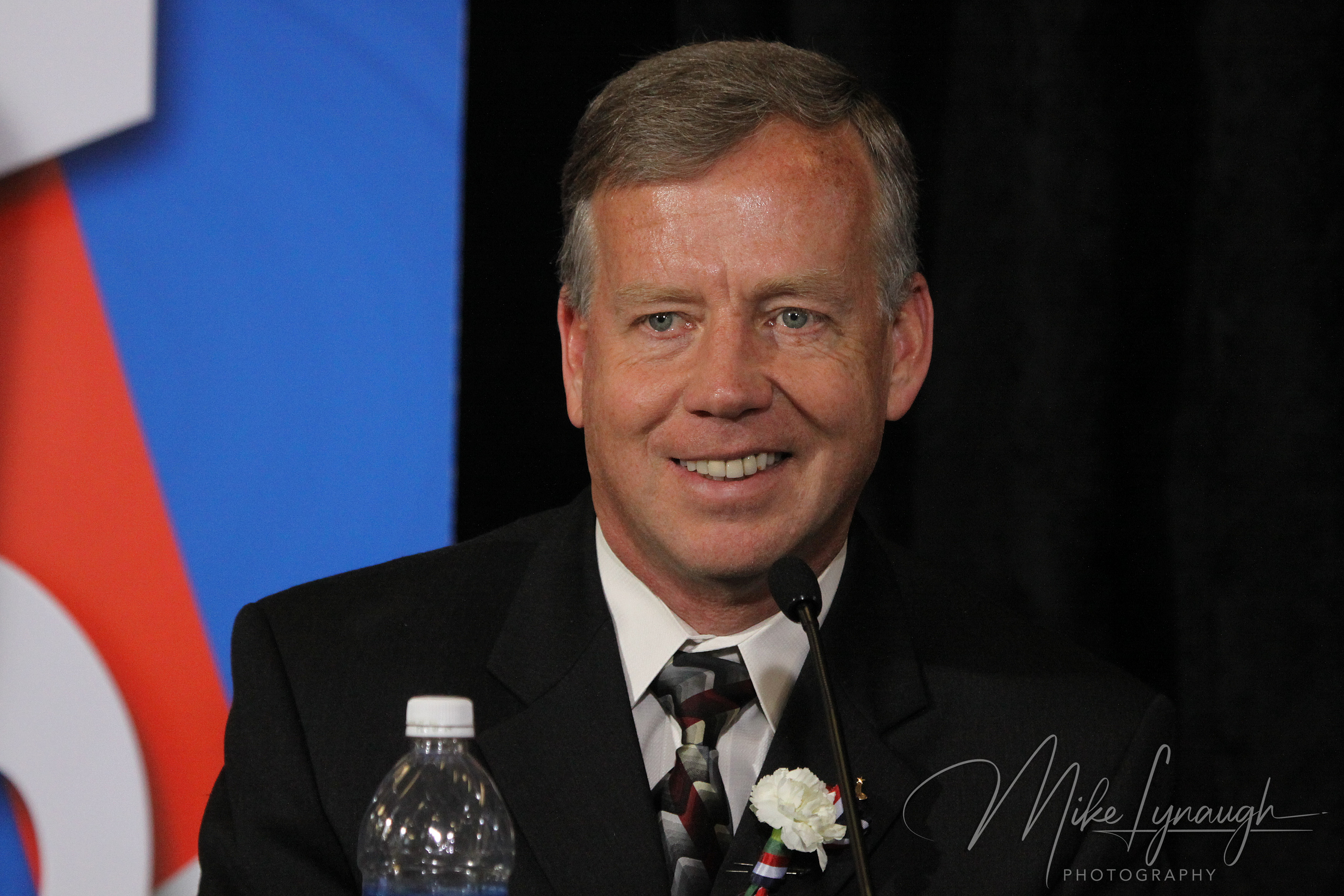 A medium close-up portrait photograph capturing former NASA astronaut Steve Lindsey—pilot of the STS-95 mission with John Glenn—smiling while addressing participants at the microphone.