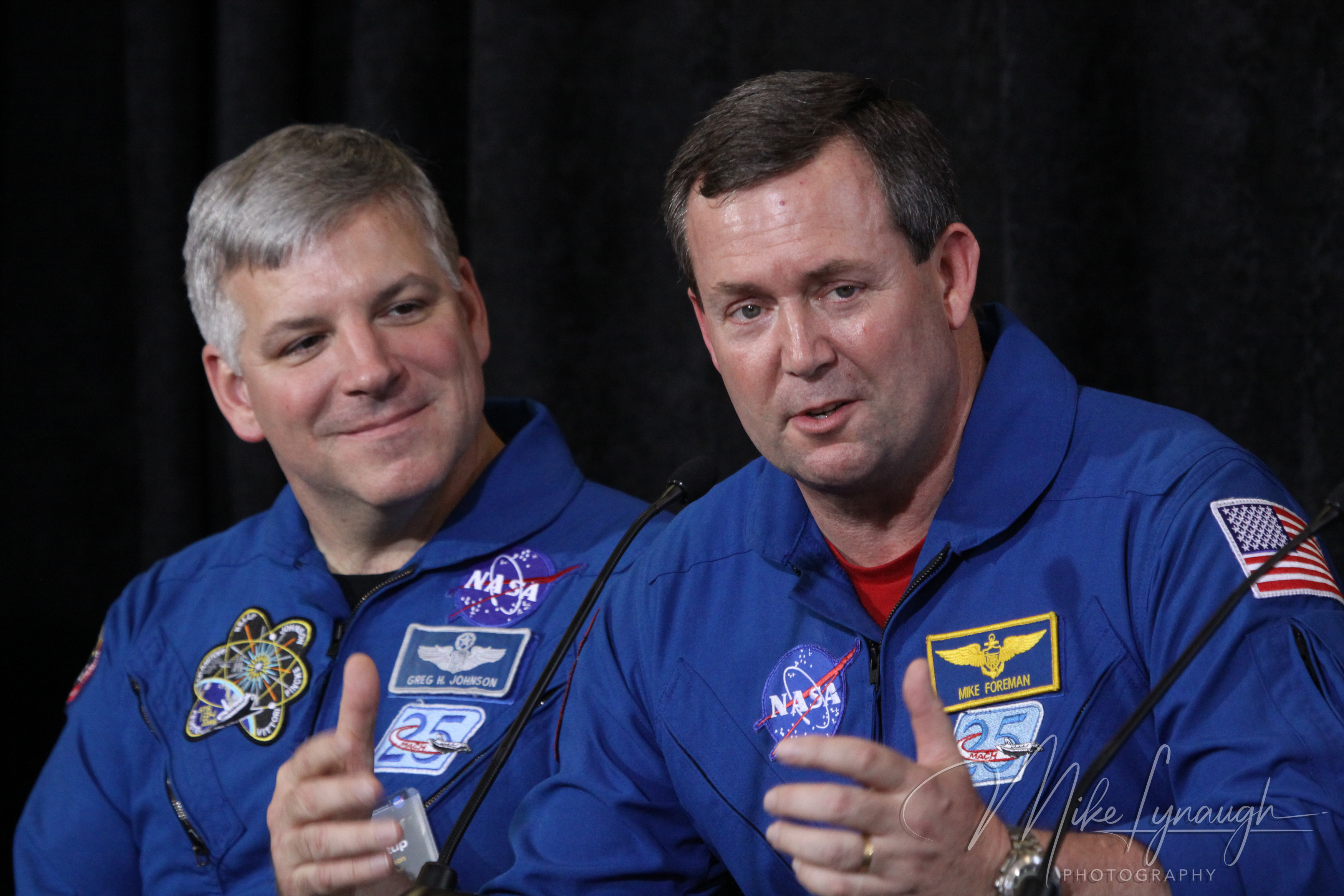 A medium close-up photograph capturing NASA astronaut Mike Foreman (right) speaking into the microphone with animated hand gestures, while fellow astronaut Gregory H.